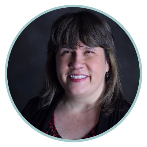 Image of Nikki Brown, PCC - A woman with shoulder-length brown hair and bangs is smiling in this headshot. She is wearing a black top with a hint of a red patterned shirt visible underneath. The background is dark, providing contrast to her light complexion. The image is enclosed within a circular border.